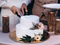 Biały tort weselny z teksturą, ozdobiony kwiatami. Para kroi tort, a obok płoną świece w drewnianych świecznikach. Trendy w prezentacji tortów weselnych to m.in. tekstury.