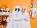 Dzieci w strojach na Halloween: jedno jako duch, drugie jako mumia. Obok zabawne dekoracje, jak pajęczyna i pluszowy potworek.