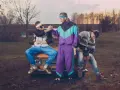 Trzech mężczyzn w strojach z lat 90. pozuje na tle starego samochodu. Jeden z nich ma na sobie fioletowy dres, drugi biało-czarną kurtkę i niebieskie spodnie, a trzeci sweter w geometryczne wzory.
