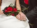 Dłonie nowożeńców splecione na tle bukietu czerwonych róż. Idealny moment, by pomyśleć, jak przygotować życzenia ślubne które zapadną w pamięć i wzruszą.