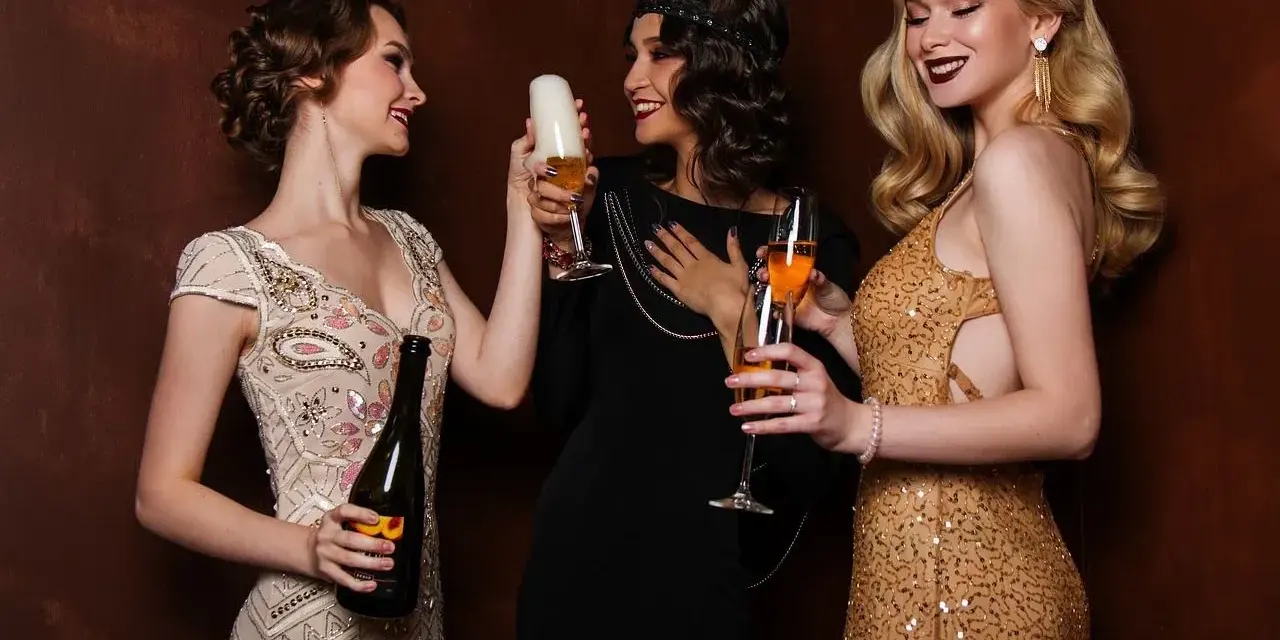 Trzy kobiety w strojach z lat 20. wznoszą toasty. Jedna trzyma butelkę szampana, druga piwo, a trzecia kieliszek z drinkiem.