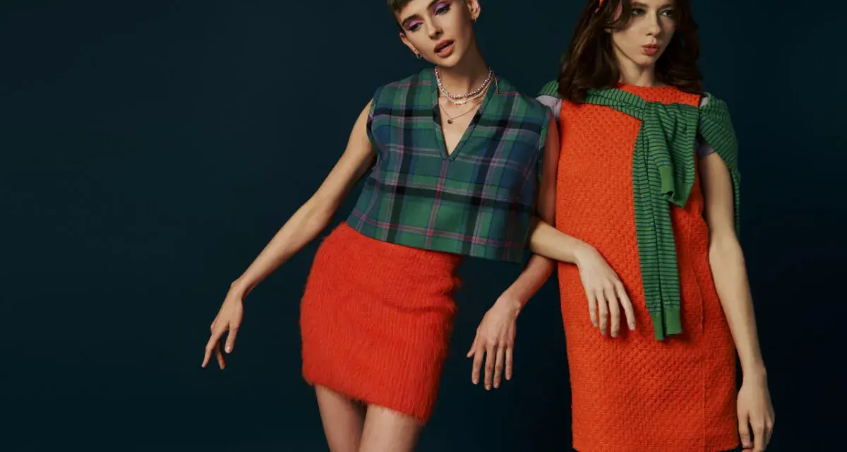 Dwie modelki w stylizacjach inspirowanych modą lat 60. Jedna w kraciastej kamizelce i pomarańczowej spódniczce, druga w pomarańczowej sukience i zielonym sweterku.