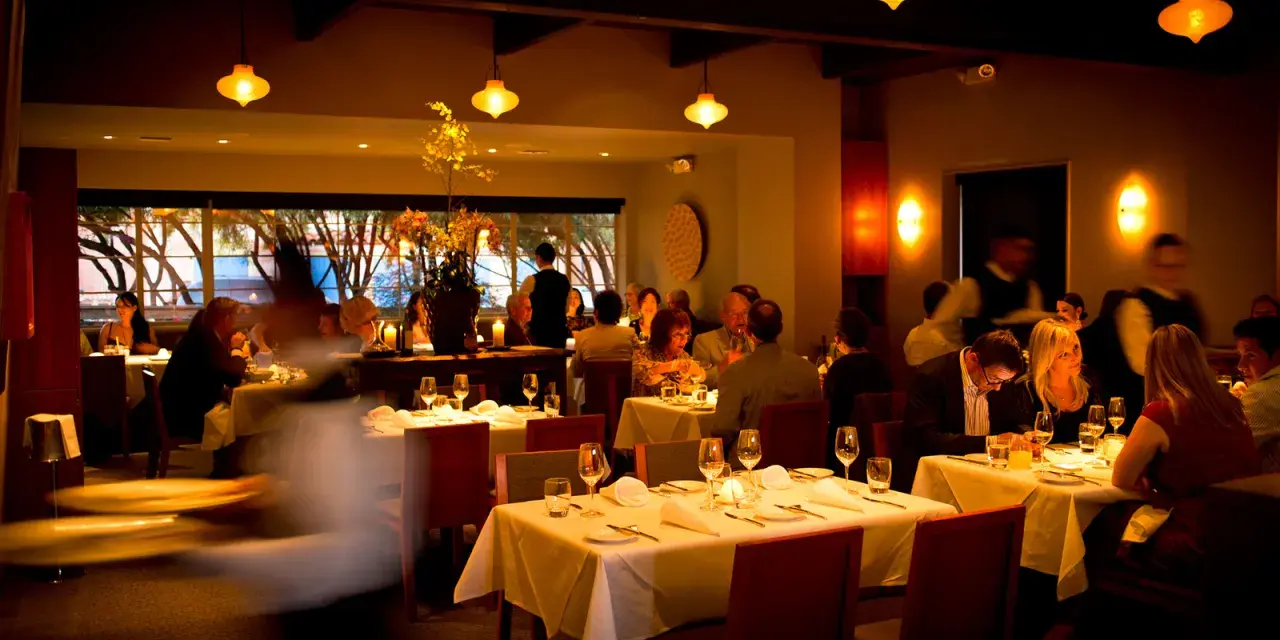 Ciemne, nastrojowe wnętrze restauracji. Goście jedzą przy stołach, kelnerzy krążą. Złe oświetlenie zdjęć jedzenia może zniechęcić gości do publikacji w social mediach.