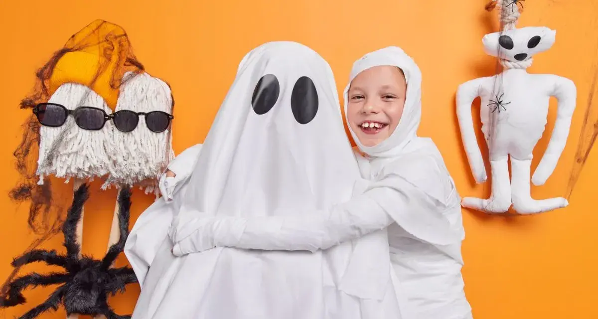 Dzieci w strojach na Halloween: jedno jako duch, drugie jako mumia. Obok zabawne dekoracje, jak pajęczyna i pluszowy potworek.