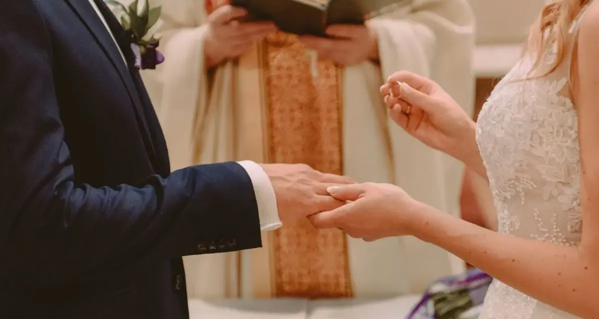 Ceremonia ślubna: pan młody w granatowym garniturze, panna młoda w białej sukni, ksiądz udziela błogosławieństwa. Czy do bycia świadkiem na ślubie trzeba mieć bierzmowanie?