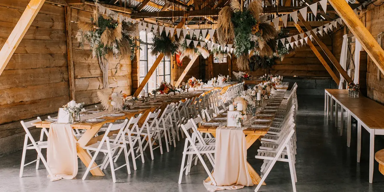 Drewniana stodoła udekorowana na przyjęcie. Długie stoły z kwiatami i świecami, wiszące dekoracje z traw. Widać różnicę między stylem boho chic a rustykalnym w dekoracjach kwiatowych.