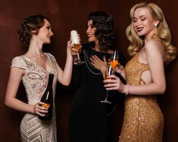 Trzy kobiety w strojach z lat 20. wznoszą toasty. Jedna trzyma butelkę szampana, druga piwo, a trzecia kieliszek z drinkiem.