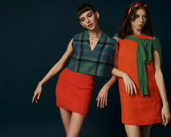 Dwie modelki w stylizacjach inspirowanych modą lat 60. Jedna w kraciastej kamizelce i pomarańczowej spódniczce, druga w pomarańczowej sukience i zielonym sweterku.