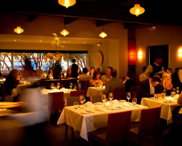 Ciemne, nastrojowe wnętrze restauracji. Goście jedzą przy stołach, kelnerzy krążą. Złe oświetlenie zdjęć jedzenia może zniechęcić gości do publikacji w social mediach.
