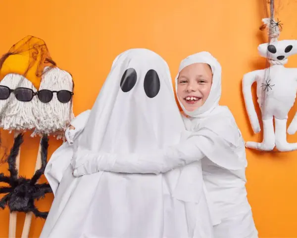 Dzieci w strojach na Halloween: jedno jako duch, drugie jako mumia. Obok zabawne dekoracje, jak pajęczyna i pluszowy potworek.