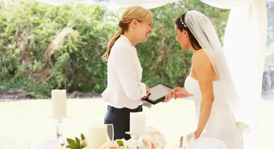 Panna młoda z welonem i wedding plannerka dyskutują przy stole. Czy warto wynająć wedding plannera, czy poradzić sobie samemu?