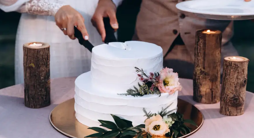Biały tort weselny z teksturą, ozdobiony kwiatami. Para kroi tort, a obok płoną świece w drewnianych świecznikach. Trendy w prezentacji tortów weselnych to m.in. tekstury.