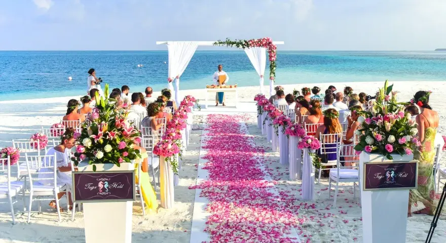 Piękna plażowa ceremonia ślubna. Kwiaty i płatki róż tworzą romantyczną ścieżkę. Goście siedzą na białych krzesłach, a ocean w tle.