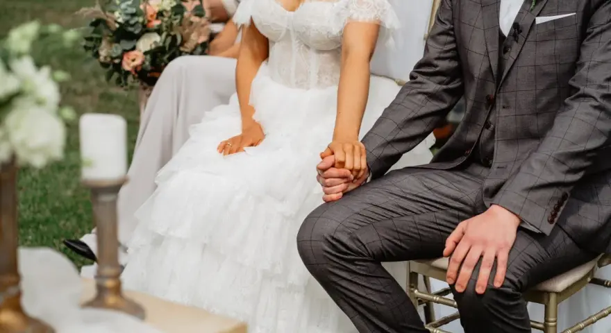 Para spowiedzi przedślubnej lub formalności w USC: panna młoda w koronkowej sukni i pan młody w garniturze trzymają się za ręce.