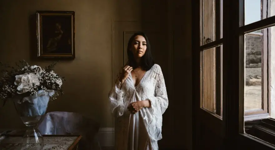Kobieta w białej sukni, obok bukiet kwiatów. Scena sugeruje przygotowania do ślubu za granicą, co wyjaśnia przyczyny popularności wesel typu "destination wedding".