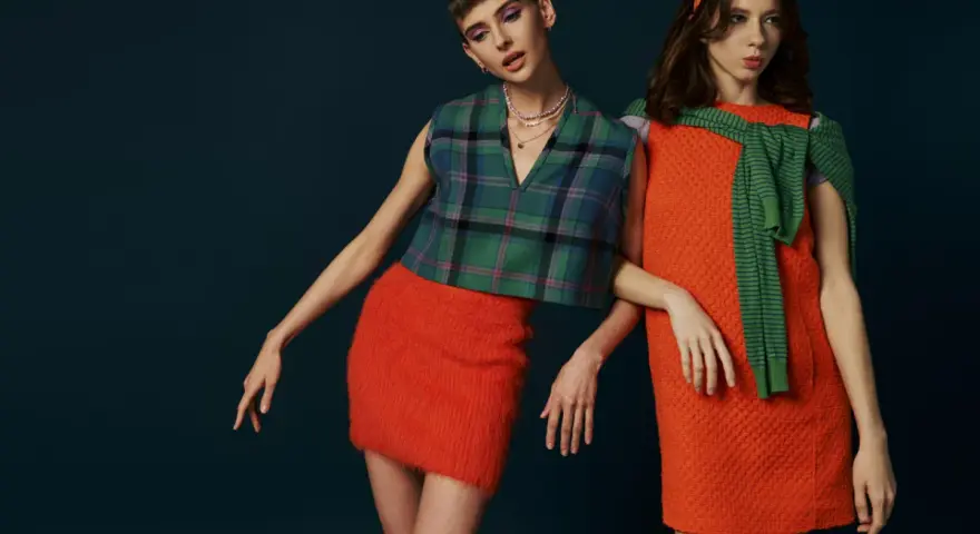 Dwie modelki w stylizacjach inspirowanych modą lat 60. Jedna w kraciastej kamizelce i pomarańczowej spódniczce, druga w pomarańczowej sukience i zielonym sweterku.