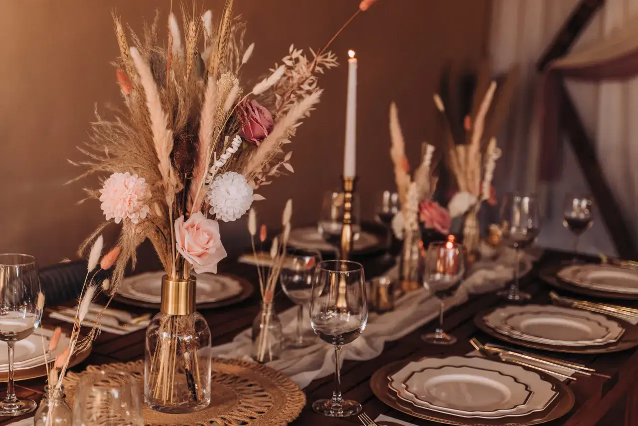 Stół udekorowany suszonymi trawami, piórami i sztucznymi kwiatami, tworząc subtelną różnicę między stylem boho chic a rustykalnym w dekoracjach kwiatowych.