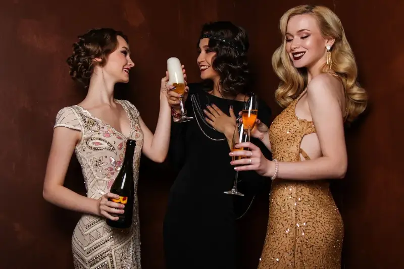Trzy kobiety w strojach z lat 20. wznoszą toasty. Jedna trzyma butelkę szampana, druga piwo, a trzecia kieliszek z drinkiem.