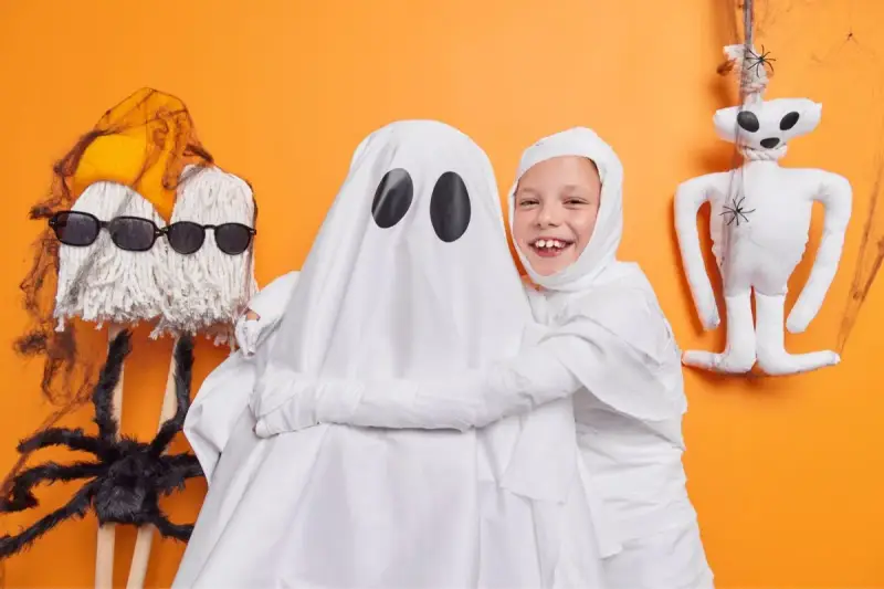 Dzieci w strojach na Halloween: jedno jako duch, drugie jako mumia. Obok zabawne dekoracje, jak pajęczyna i pluszowy potworek.